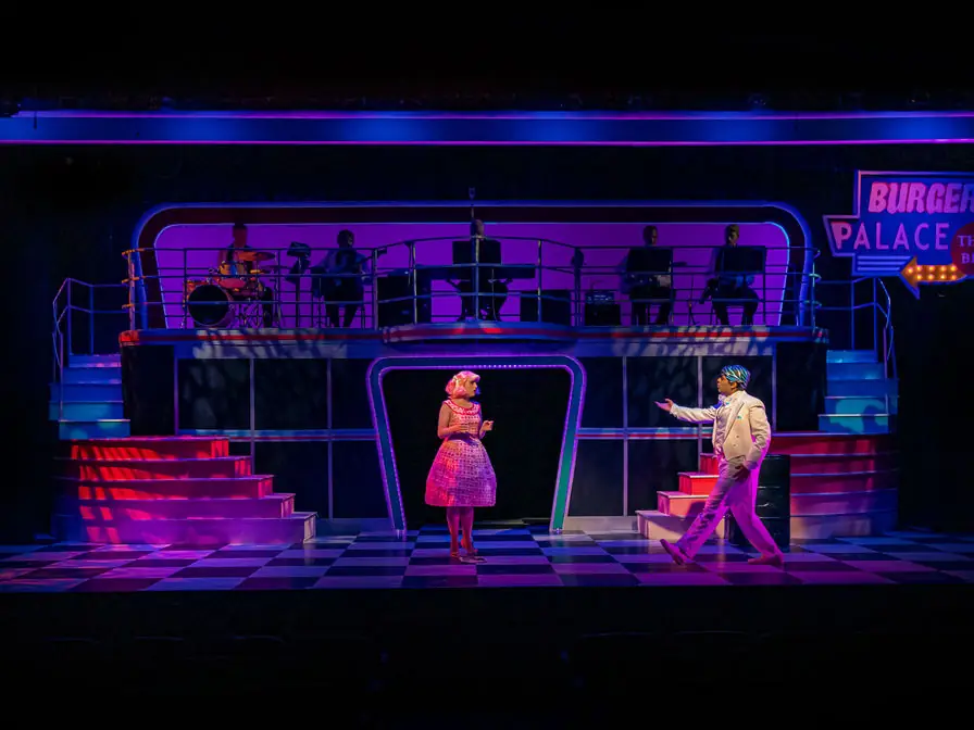 Wide production photo from Grease showing the full company dancing in front of Gretchen Ugalde's neon diner-inspired set.